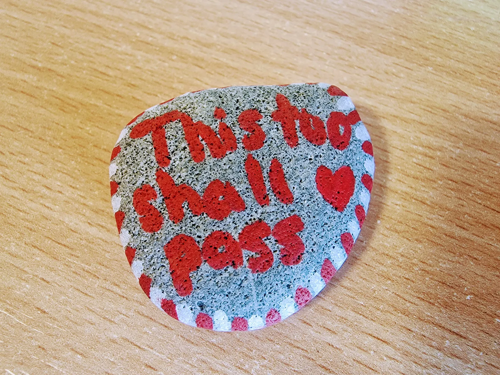 A photo of a small rock with red writing saying 'this too shall pass', with white and red stripes around the outside