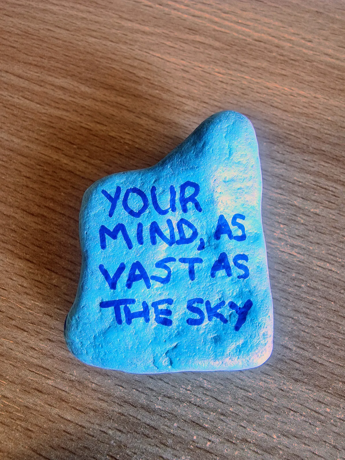 A photo of a small roughly square-shaped rock painted in light blue, with dark blue text saying 'your mind, as vast as the sky'.